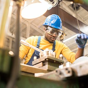 Serious busy young black factory engineer in hardhat and safety goggles examining milling lathe and repairing it while working at production plant