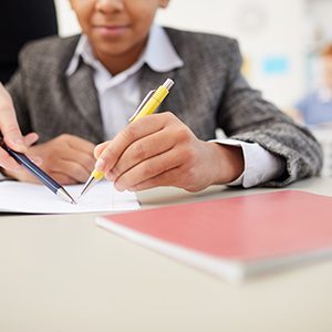 Close-up of African boy sitting at desk and working in his notebook with teacher pointing at the mistake during a lesson