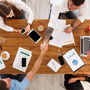 Business meeting. Busy people work in office, top view of wood table with mobile phones, laptop, tablet and documents papers with diagram. Men and women team handshake, have contract conclusion.