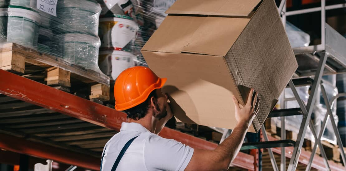Worker carrying a box while standing on a ladder, illustrating workplace ergonomics and manual handling risks.